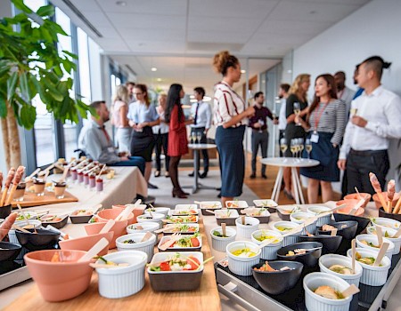 A group of people socializing at a reception or party, with a buffet table in the foreground featuring various small dishes and appetizers.