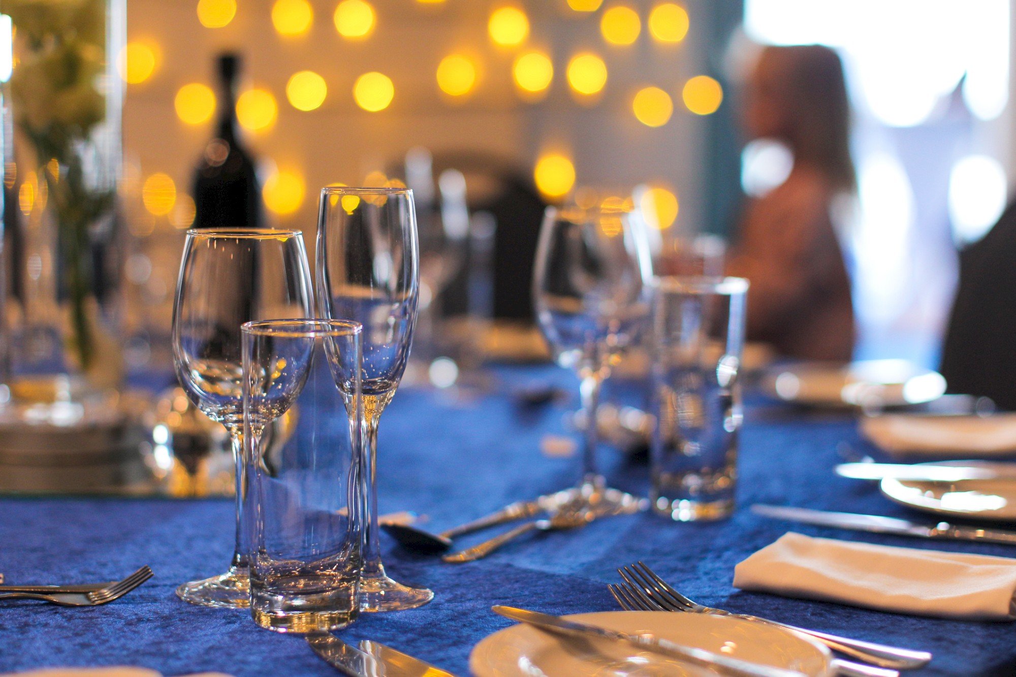 A table is elegantly set with glassware and cutlery on a blue tablecloth, with soft bokeh lights in the background.