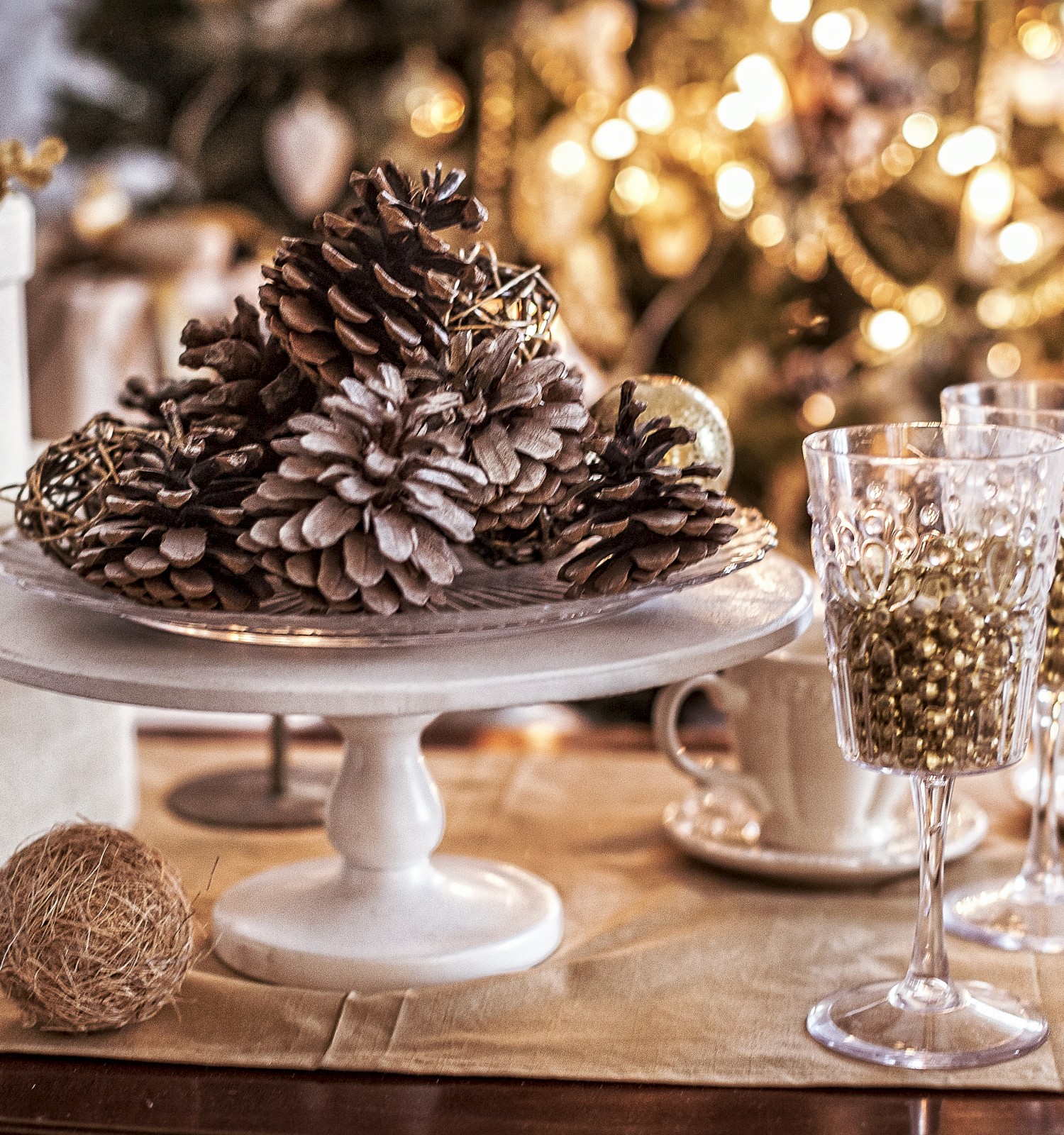 Festive table decor with pine cones on a stand, wine glasses, a string ball, and a blurred Christmas tree background with lights.