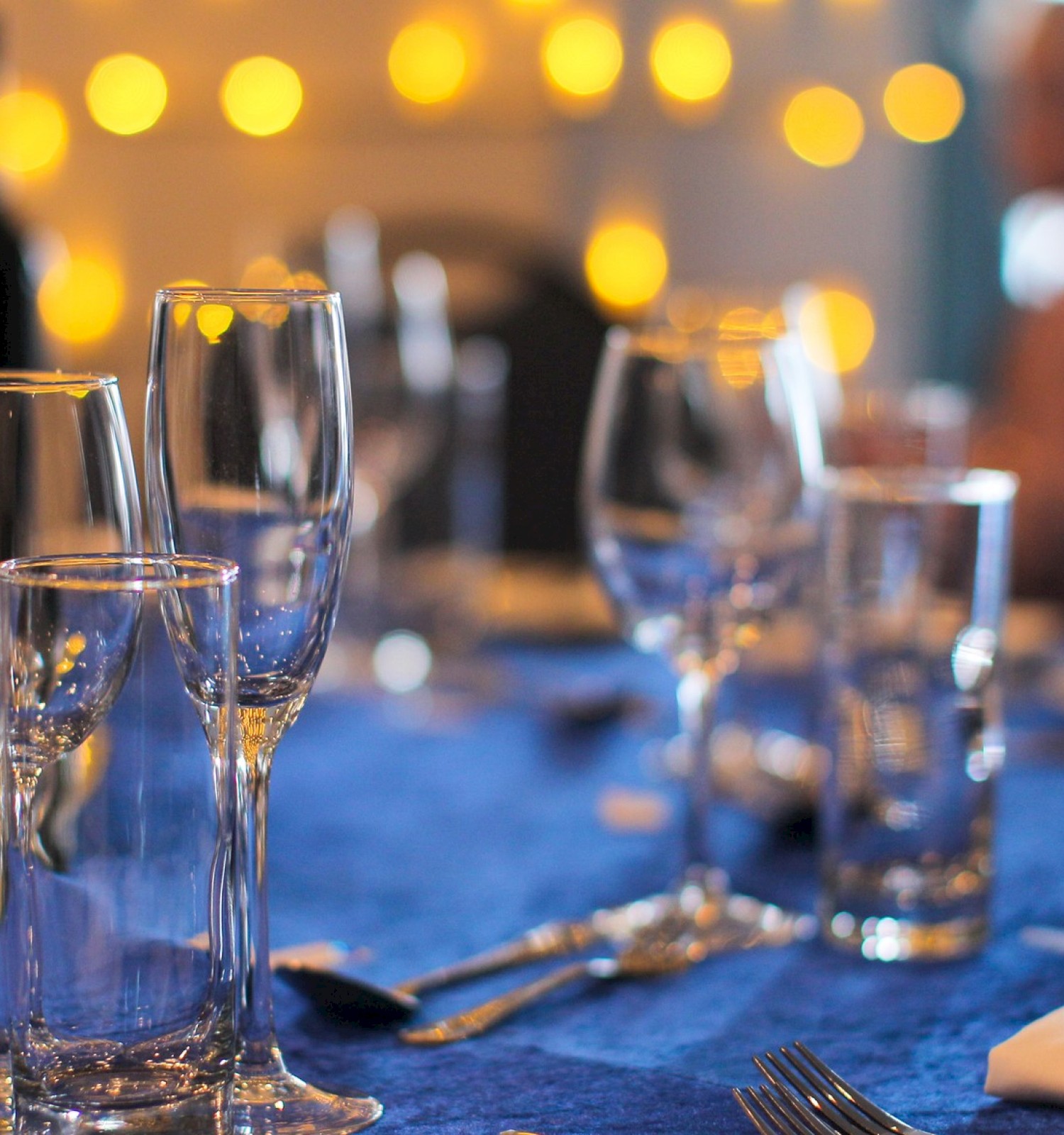 A table set for a formal event with glasses, plates, and cutlery on a blue tablecloth; blurred lights create a warm, festive atmosphere.