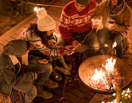 Four people in cozy sweaters roast marshmallows over a firepit on a wooden deck, surrounded by string lights, enjoying a warm evening.