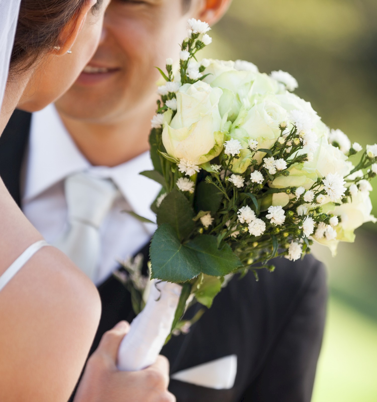 A bride holds a bouquet of white flowers, facing her groom in a suit with a tie, outdoors.