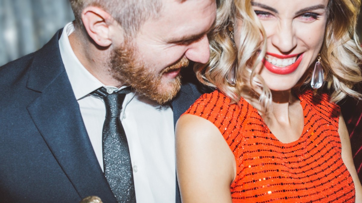 A smiling couple dressed formally; the woman in a red dress, the man in a suit holding a bottle, enjoying a lively moment together.