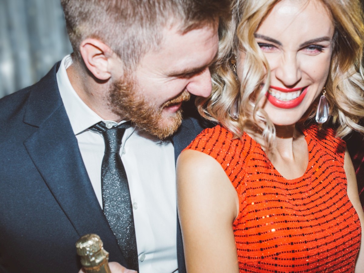 A smiling couple dressed formally; the woman in a red dress, the man in a suit holding a bottle, enjoying a lively moment together.