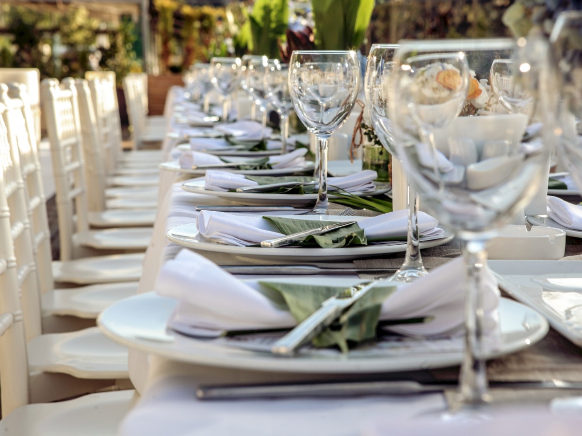 A long, elegantly set dining table with white tableware, folded napkins, and wine glasses, surrounded by white chairs in an outdoor setting.