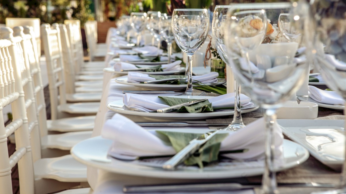 A long, elegantly set dining table with white tableware, folded napkins, and wine glasses, surrounded by white chairs in an outdoor setting.