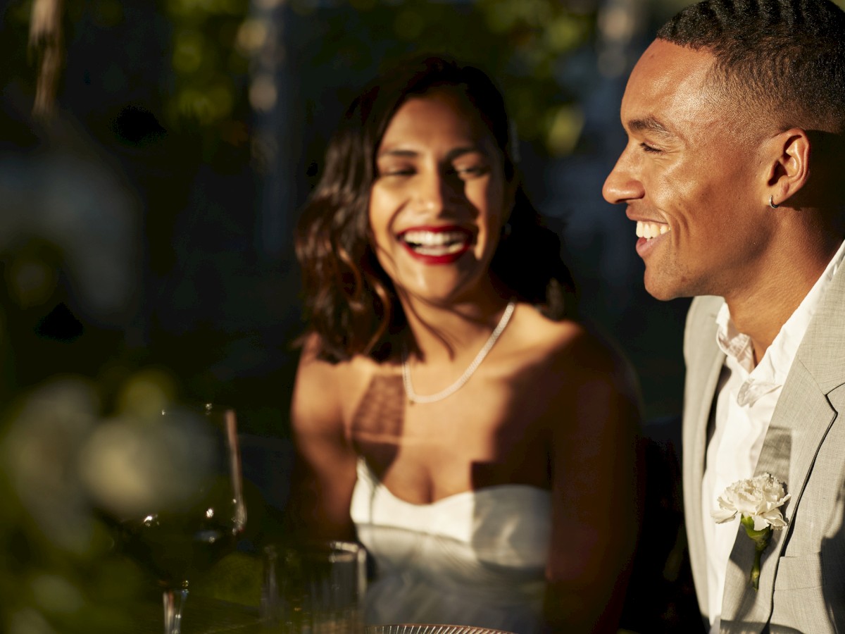 A couple is smiling and sitting together at an elegantly set table, wearing formal attire, with a warm ambiance in the background.