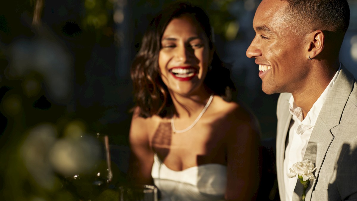 A couple is smiling and sitting together at an elegantly set table, wearing formal attire, with a warm ambiance in the background.