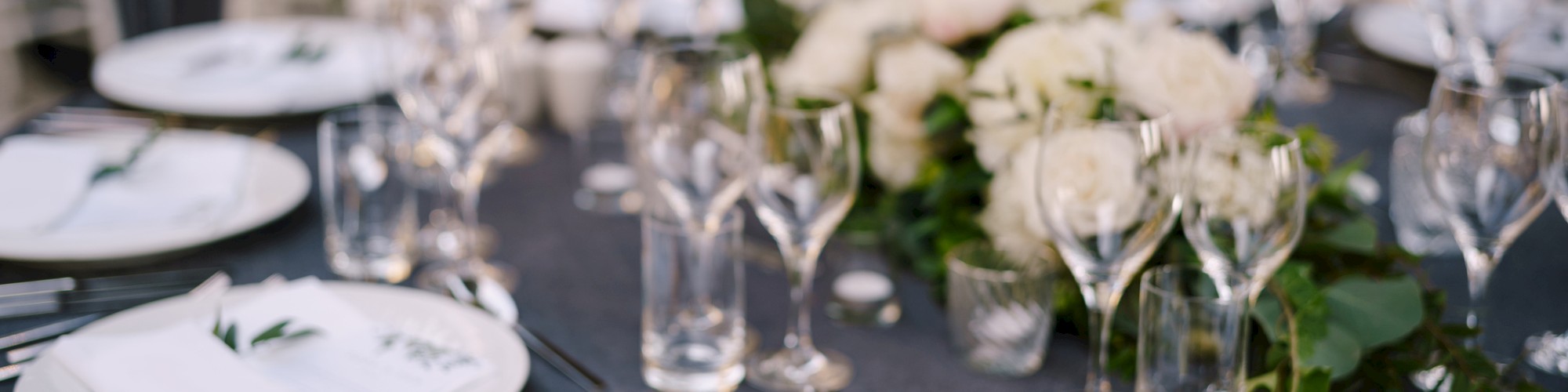 A table is elegantly set with white plates, glassware, and floral centerpieces, surrounded by white chairs, creating a formal dining ambiance.