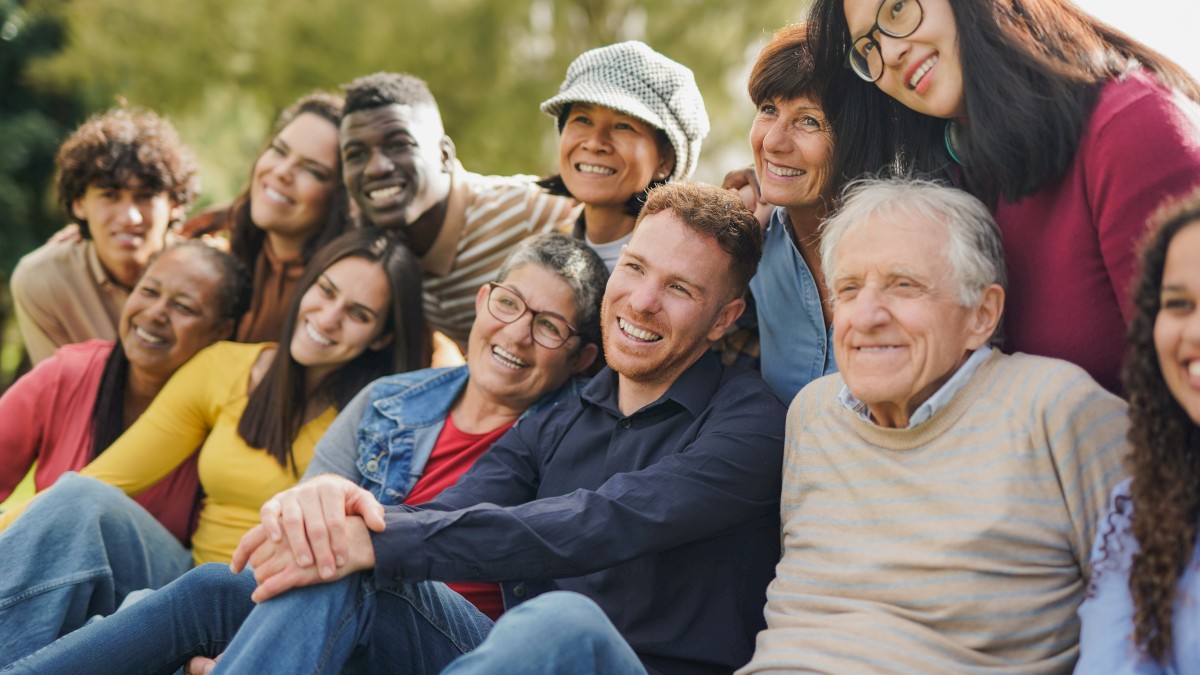 A diverse group of people sitting close together outdoors, smiling and looking happy, with greenery in the background.