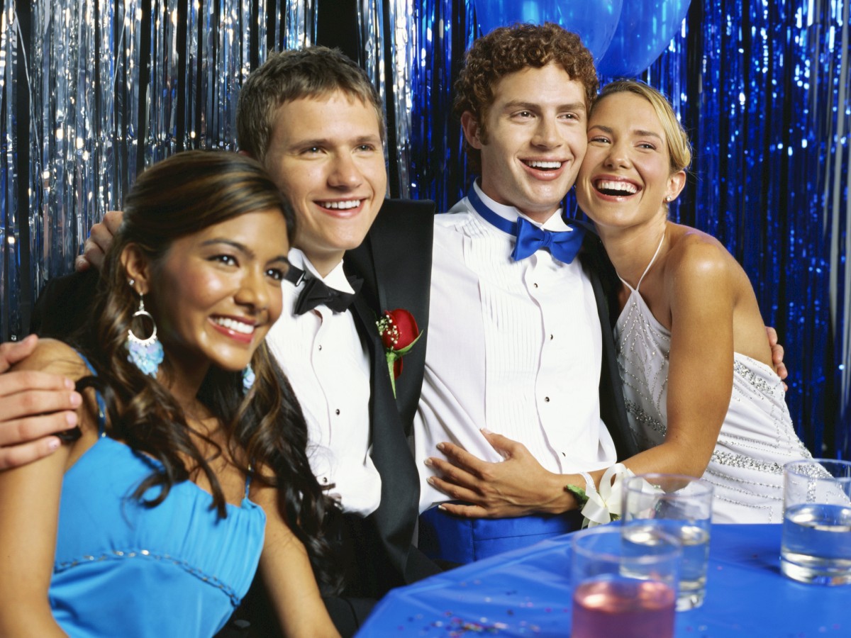 A group of four people dressed formally, smiling, at a party with a blue and silver backdrop and drinks on the table.