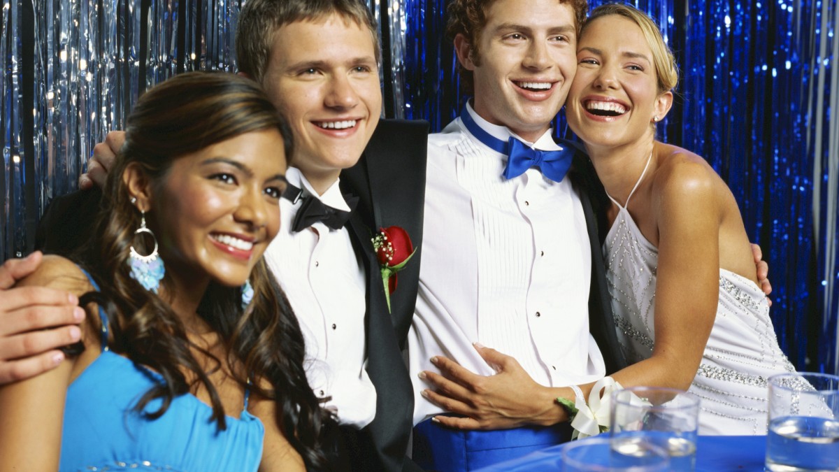 A group of four people dressed formally, smiling, at a party with a blue and silver backdrop and drinks on the table.