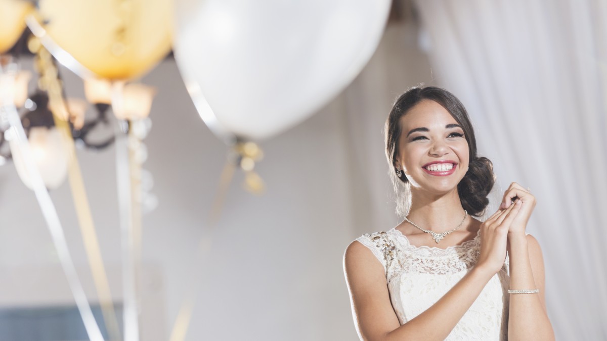 A smiling woman in a white dress stands near balloons at a celebration, looking joyful and happy.