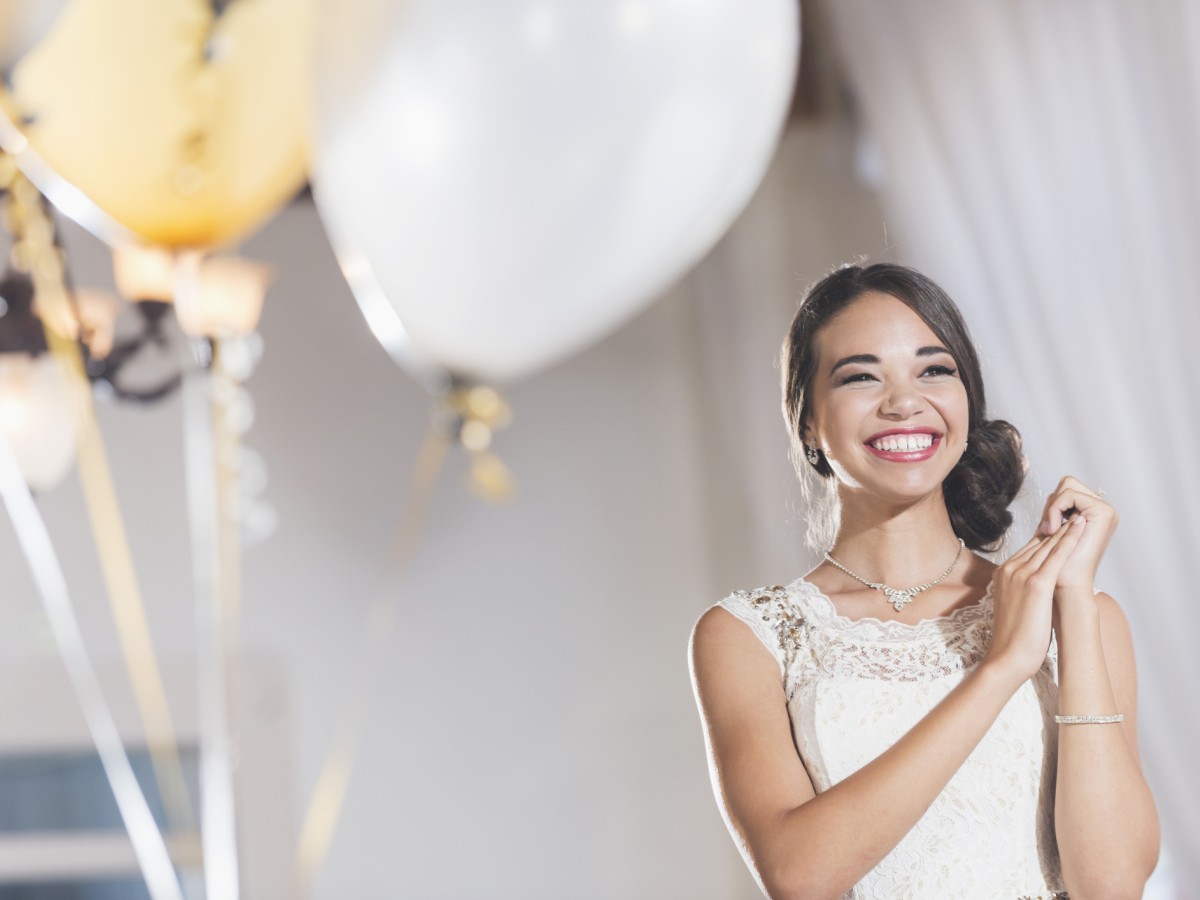 A smiling woman in a white dress stands near balloons at a celebration, looking joyful and happy.