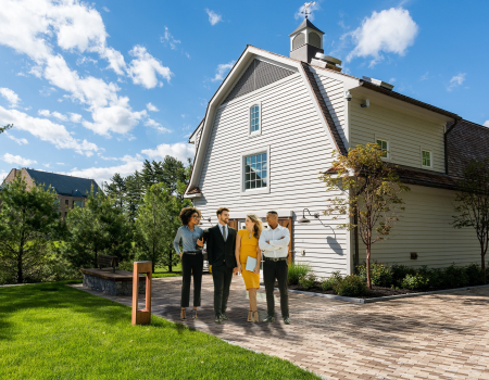 A group of four people stand in front of a large, white, two-story house on a sunny day, surrounded by green trees and a brick driveway.