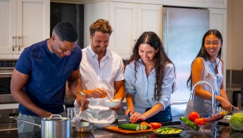 Four people are preparing food together in a kitchen, surrounded by vegetables and cookware, enjoying the cooking process.