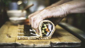 Hands are rolling sushi using a bamboo mat, with visible vegetables and rice, on a wooden surface.