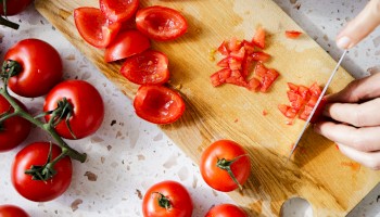 Fresh tomatoes are being sliced on a wooden cutting board, surrounded by whole tomatoes on a terrazzo countertop.