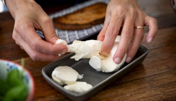 Hands preparing slices of fresh mozzarella on a dark rectangular dish, with bread and greens nearby on a wooden table.