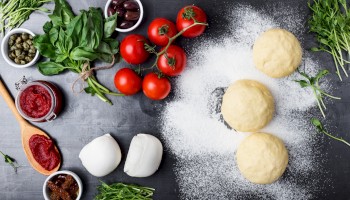 Pizza ingredients: dough balls, tomatoes, mozzarella, tomato sauce, basil, olives, capers, and sun-dried tomatoes on a dark surface.