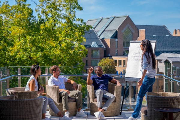 A group of people are having a meeting outdoors on a terrace with a flipchart, surrounded by trees and buildings under a clear sky.