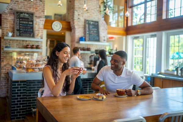 A couple is enjoying coffee at a cafe with pastries on the table, sitting at a wooden table in a cozy, brick-walled setting.