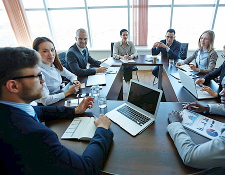 A group of people in business attire having a meeting around a conference table with laptops and documents in a bright office room.