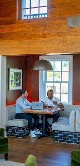 The image shows a cozy meeting space with people chatting, seated in comfortable chairs and a booth, surrounded by warm wooden decor.