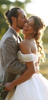 A couple in wedding attire is embracing outdoors, with the groom kissing the bride on the cheek as she holds a bouquet.